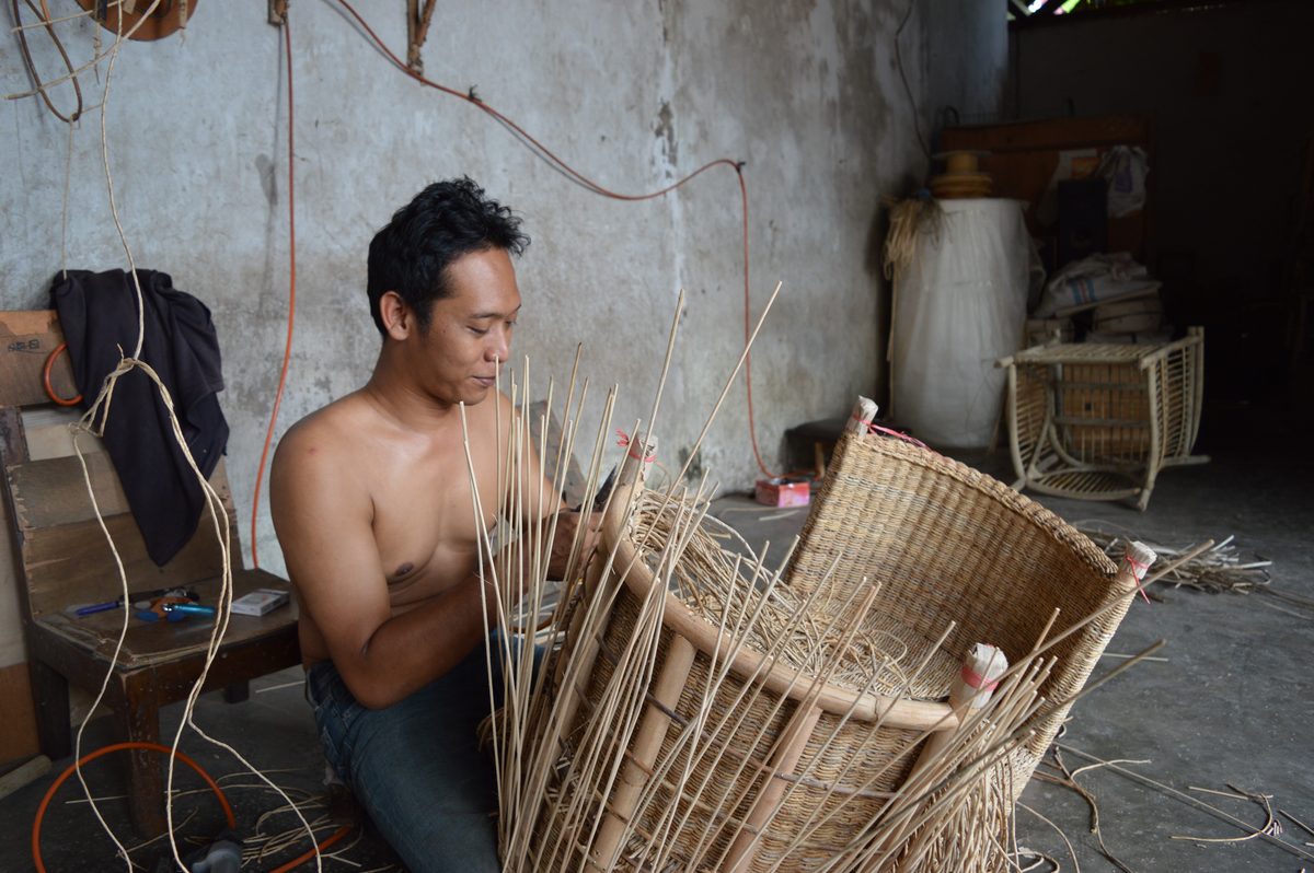A maker weaving rattan around a chair frame in a workshop in Java